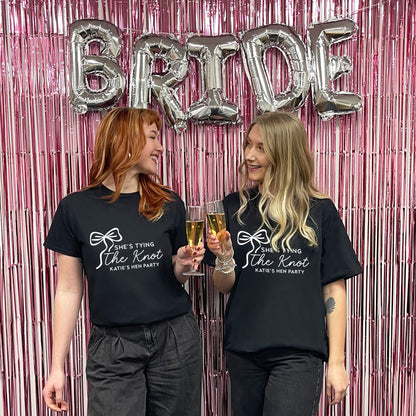 Two women wearing black t-shirts with 'She's Tying the Knot' text, holding drinks against a pink 'BRIDE' balloon backdrop.