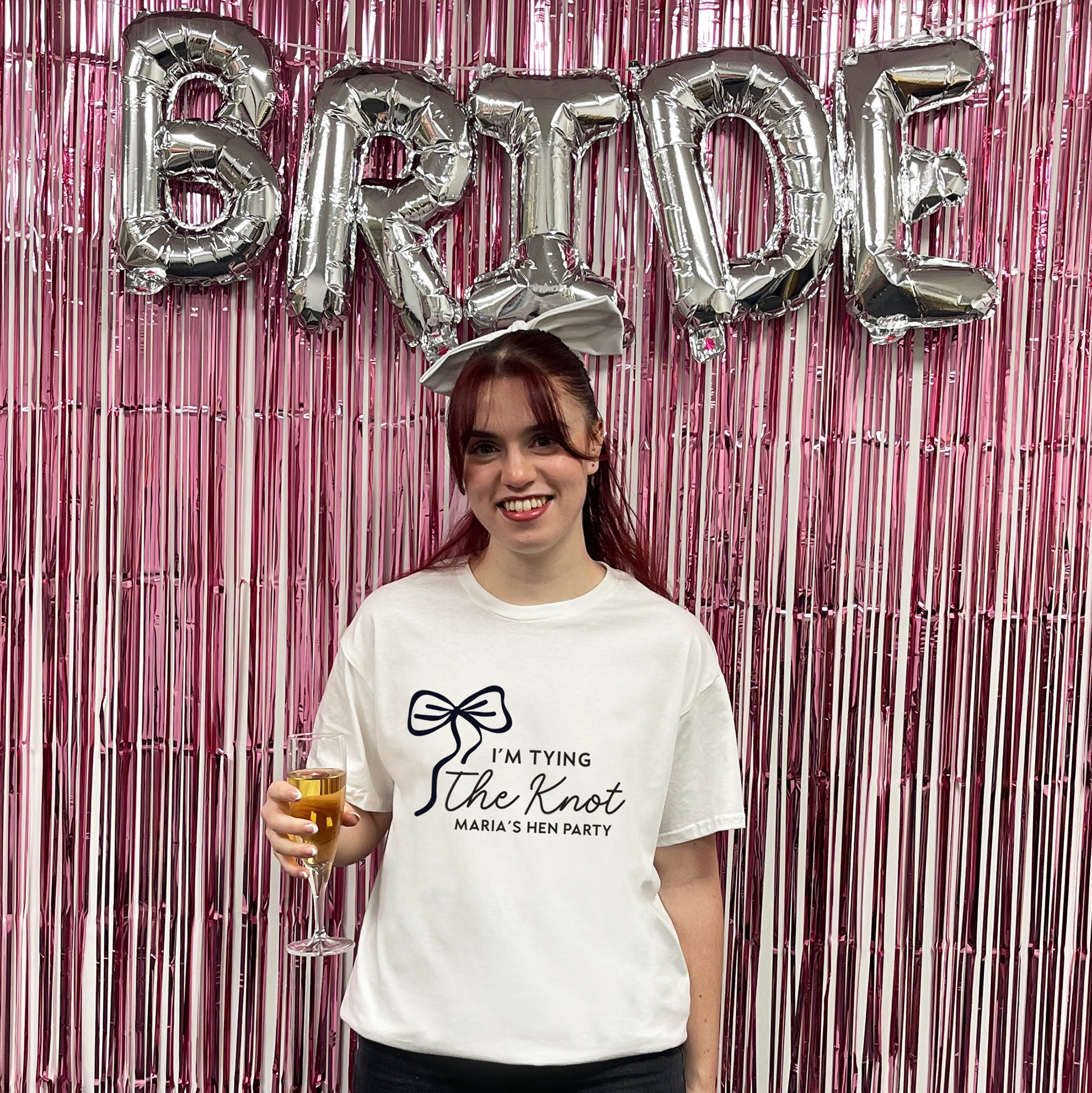 Woman holding a glass of champagne with 'BRIDE' balloons and a t-shirt with text in front of a pink fringe curtain.