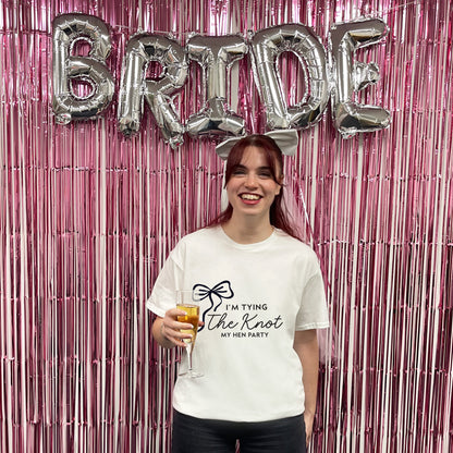 Woman holding a drink with 'Bride' balloons above her against a pink tinsel curtain.
