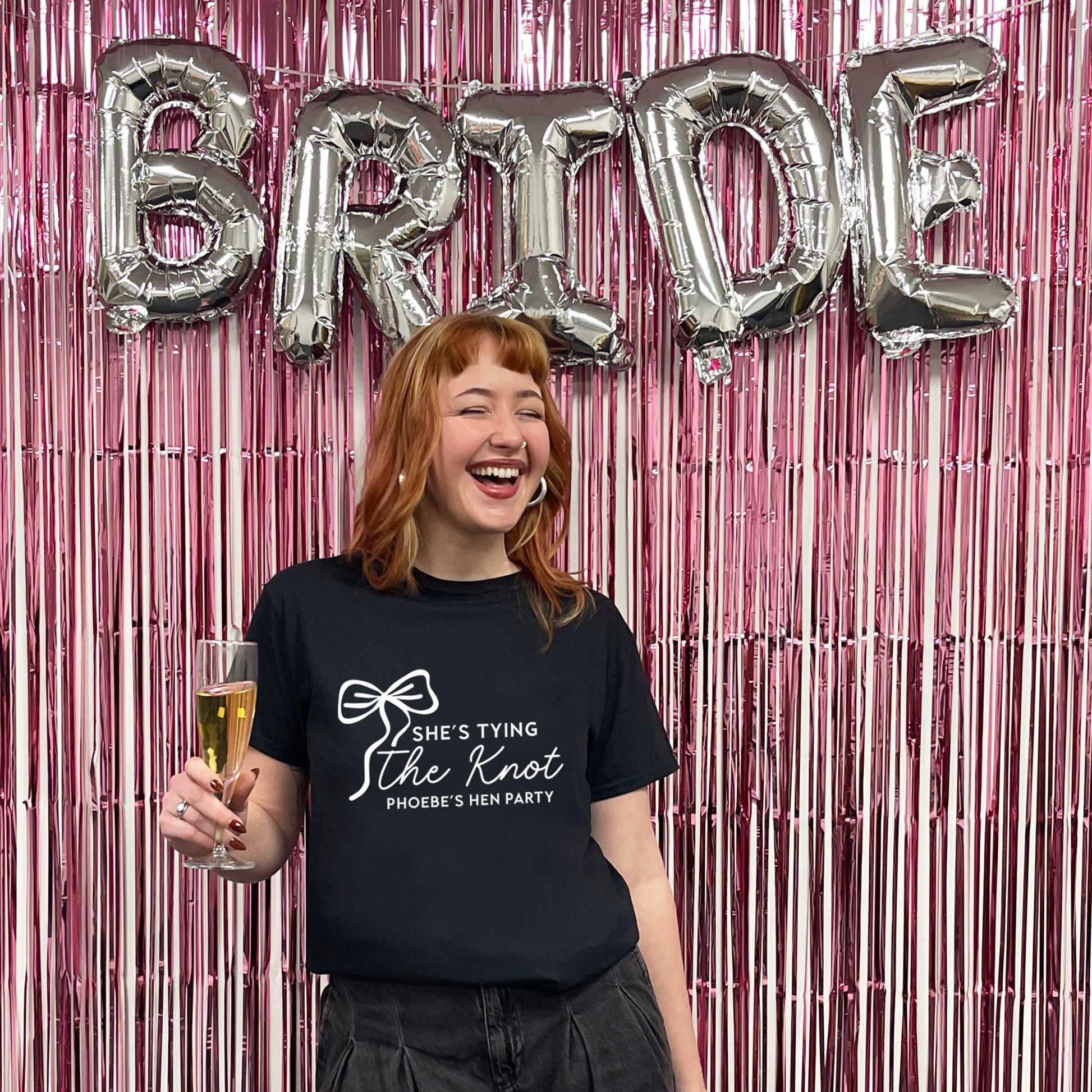 Woman wearing a black t-shirt with text, holding a glass, in front of 'BRIDE' balloons and pink streamers.