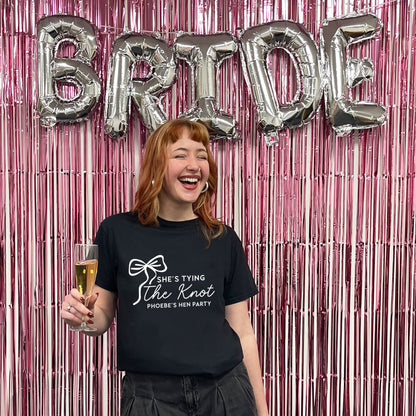 Woman wearing a black t-shirt with text, holding a glass, in front of 'BRIDE' balloons and pink streamers.