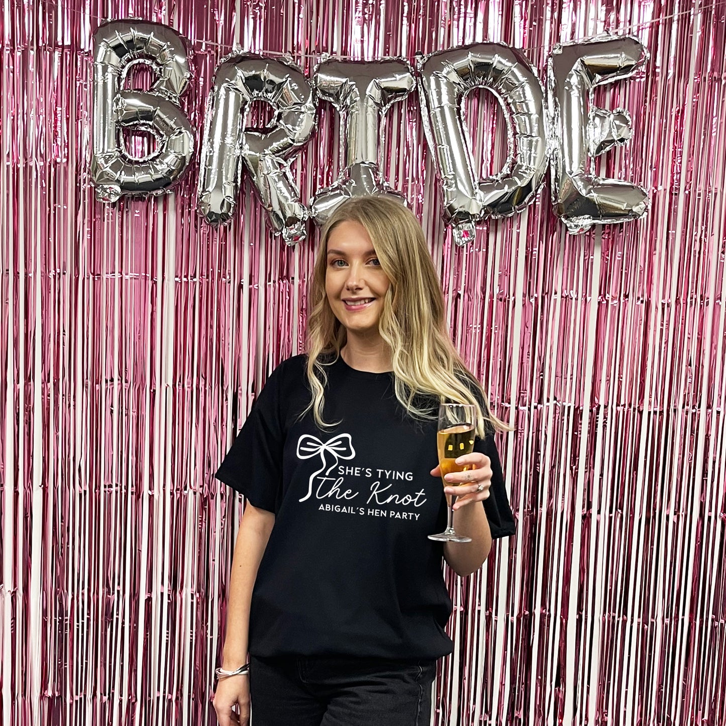 Woman holding a glass of champagne in front of 'BRIDE' balloons and pink fringe curtain.