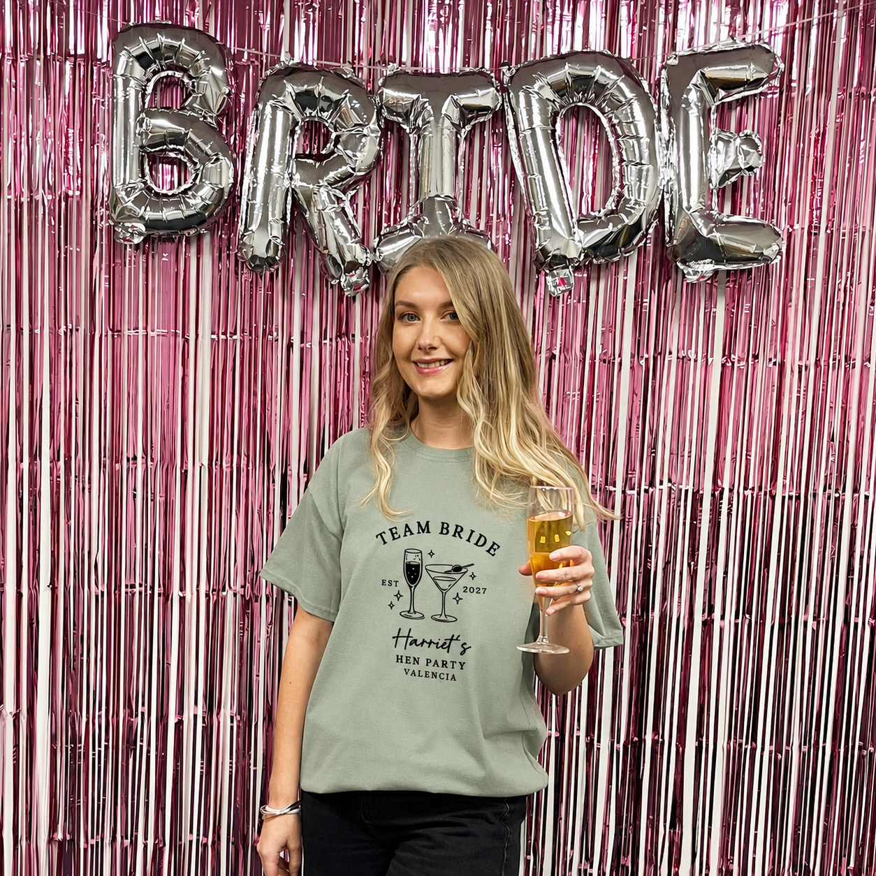 A woman stands in front of a pink metallic tinsel backdrop with large silver foil balloons spelling BRIDE. She is wearing a sage green graphic T-shirt that reads 'TEAM BRIDE' and 'Handy's HEN PARTY CALIFORNIA' with a cocktail illustration, and she holds a glass of champagne.