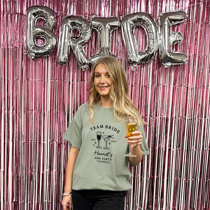 A woman stands in front of a pink metallic tinsel backdrop with large silver foil balloons spelling BRIDE. She is wearing a sage green graphic T-shirt that reads 'TEAM BRIDE' and 'Handy's HEN PARTY CALIFORNIA' with a cocktail illustration, and she holds a glass of champagne.