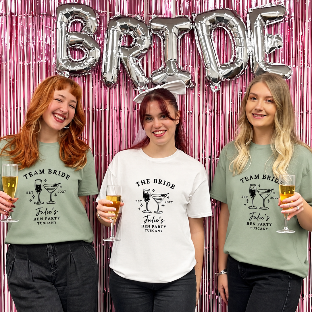 Three women posing together in front of a pink metallic tinsel backdrop with silver 'BRIDE' balloons. They are wearing white and sage green 'Team Bride' graphic t-shirts and holding champagne glasses.