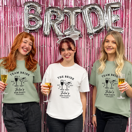 Three women posing together in front of a pink metallic tinsel backdrop with silver 'BRIDE' balloons. They are wearing white and sage green 'Team Bride' graphic t-shirts and holding champagne glasses.
