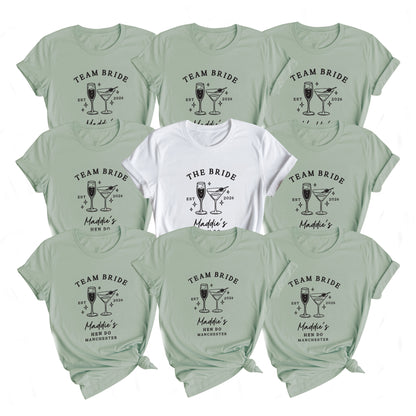 A flat lay of ten short-sleeve graphic T-shirts arranged in a grid on a white background. Nine shirts are a pale sage green with a black 'Team Bride' cocktail graphic, and the center shirt is white with a black 'The Bride' cocktail graphic.