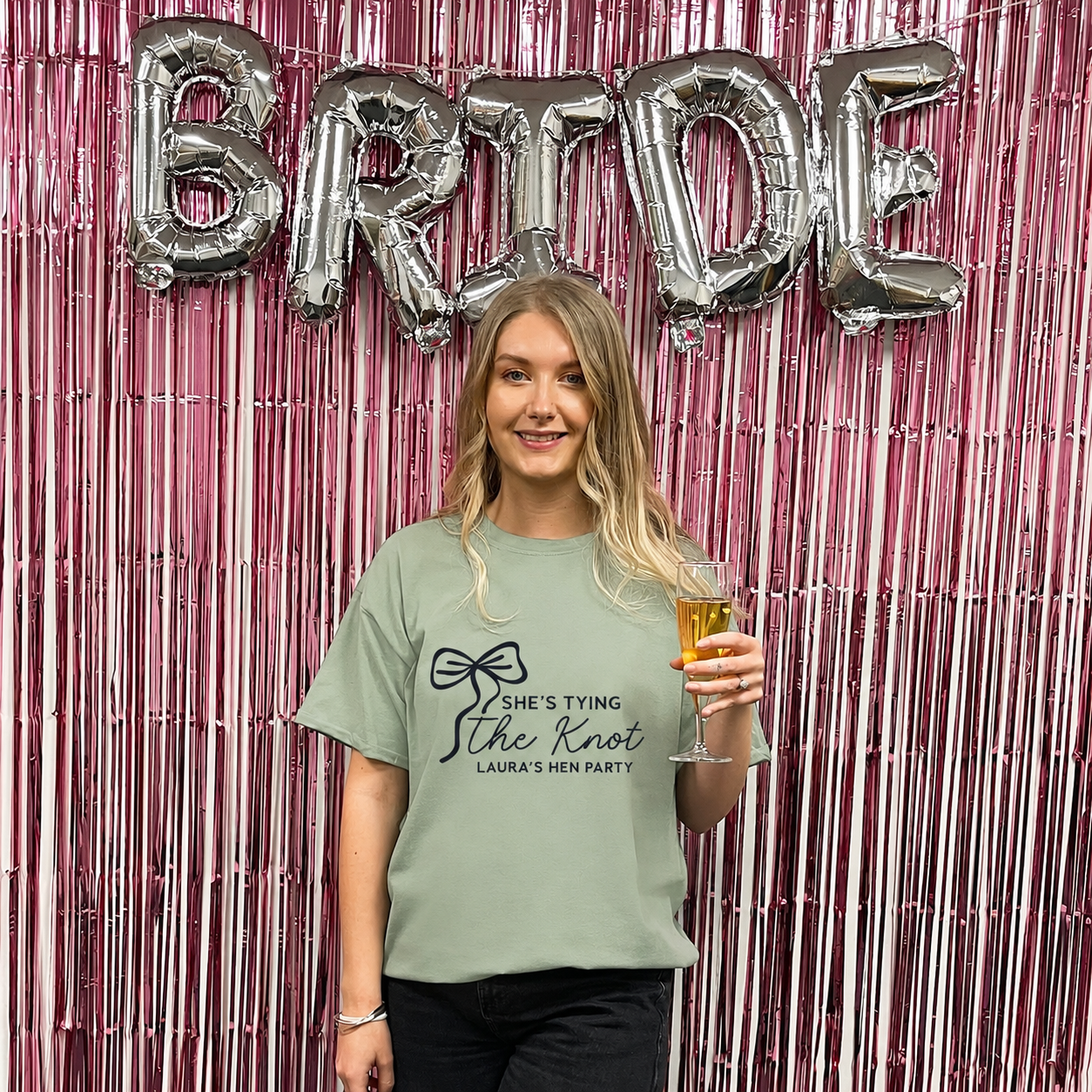Woman wearing a t-shirt with 'She's Tying the Knot' text, holding a glass, in front of 'BRIDE' balloons.