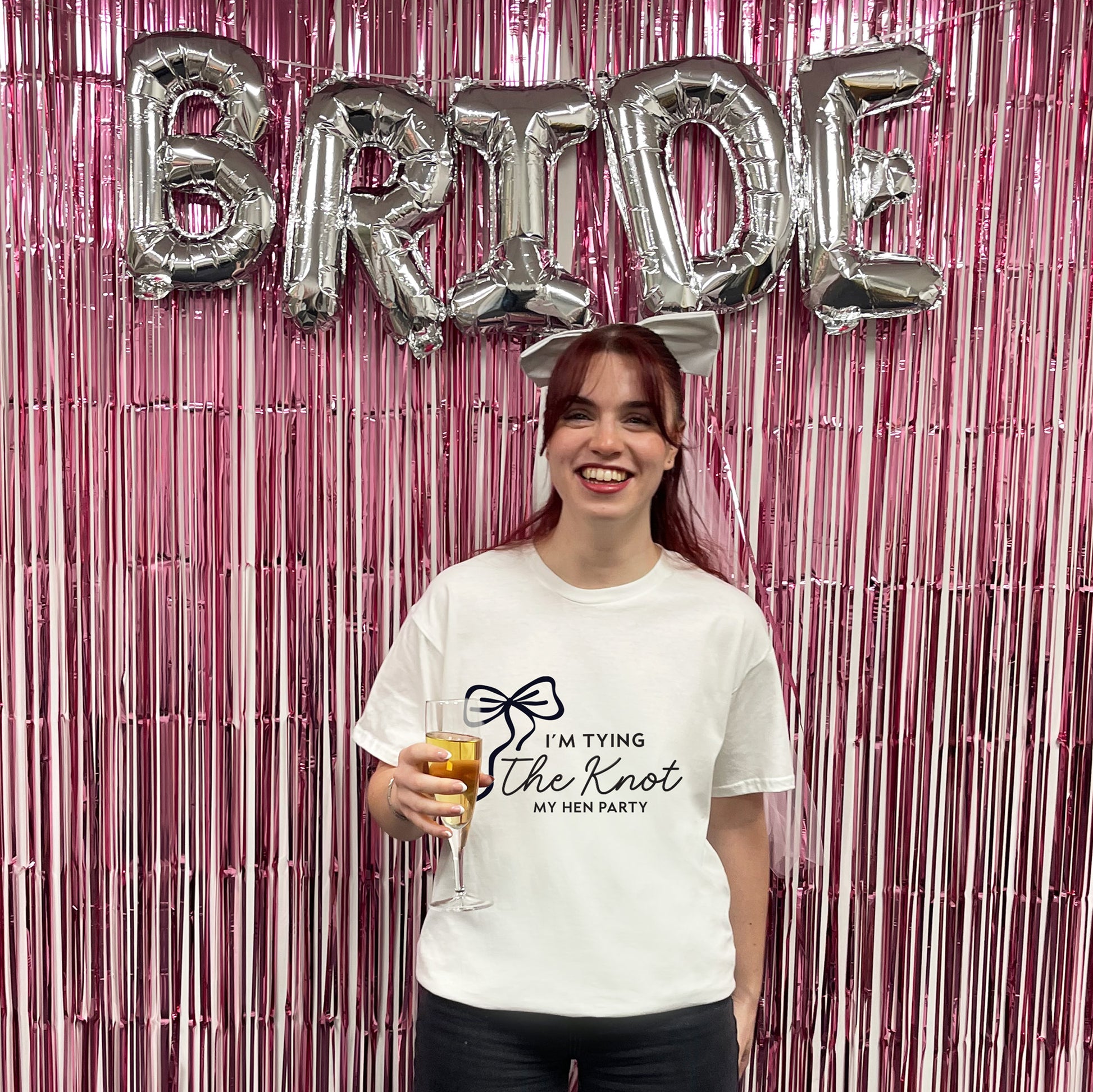 Woman holding a drink with 'Bride' balloons above her against a pink tinsel curtain.