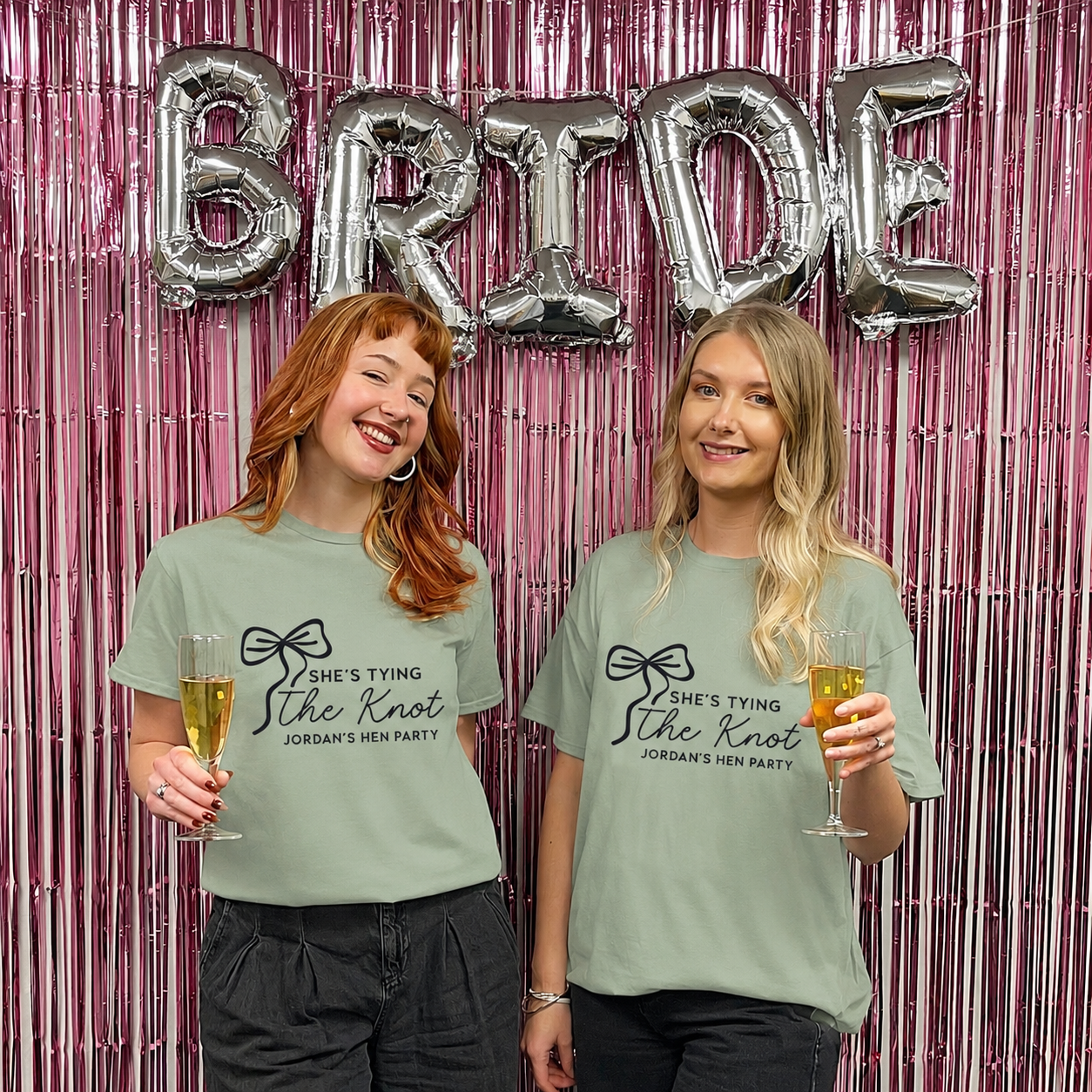 Two women wearing matching t-shirts with 'She's tying the knot' text, holding drinks in front of a 'BRIDE' balloon backdrop.