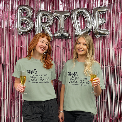 Two women wearing matching t-shirts with 'She's tying the knot' text, holding drinks in front of a 'BRIDE' balloon backdrop.