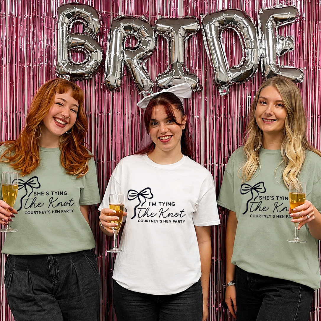 Three women wearing matching t-shirts with a 'Bride' sign and pink decorative curtain.