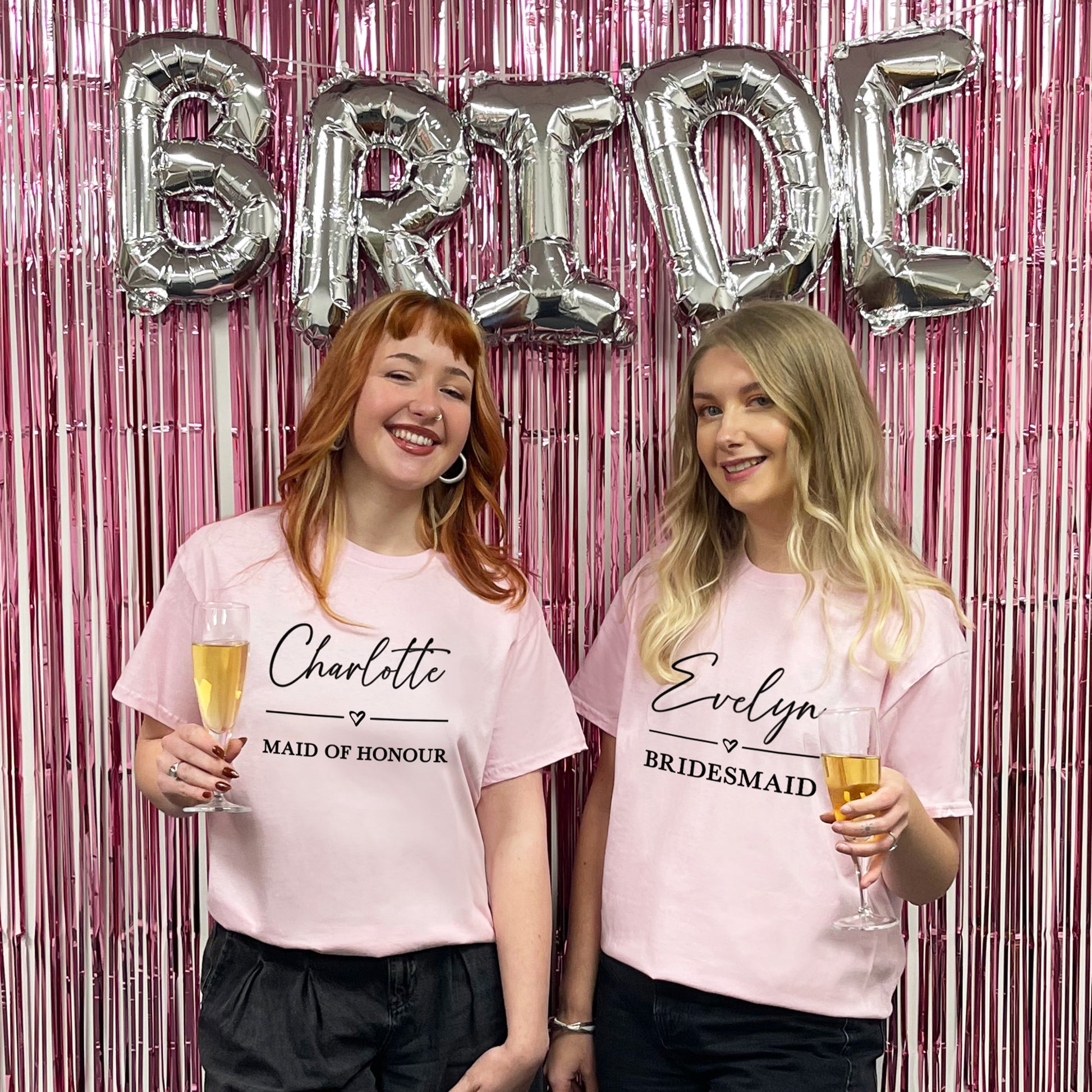 Two women in pink t-shirts with 'Bride', 'Maid of Honour', and 'Bridesmaid' text, holding drinks against a pink and silver 'Bride' balloon backdrop.