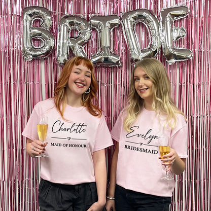 Two women in pink t-shirts with 'Bride', 'Maid of Honour', and 'Bridesmaid' text, holding drinks against a pink and silver 'Bride' balloon backdrop.