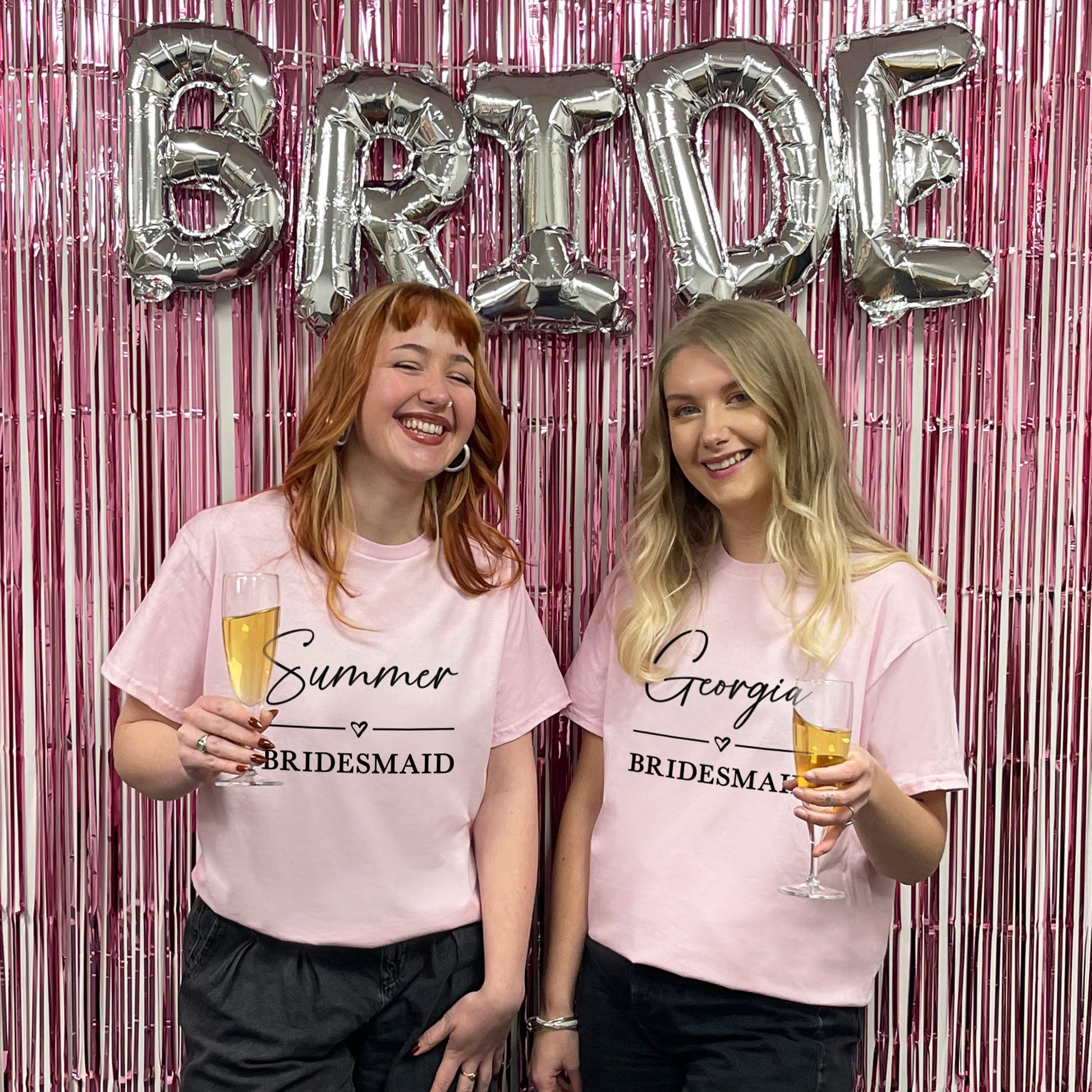 Two women in pink 'Bridesmaid' shirts holding drinks in front of a 'BRIDE' balloon and pink tinsel curtain.