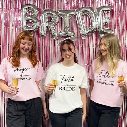 Three women in t-shirts with 'Bride' and 'Bridesmaid' text, celebrating a wedding with pink and silver decorations.