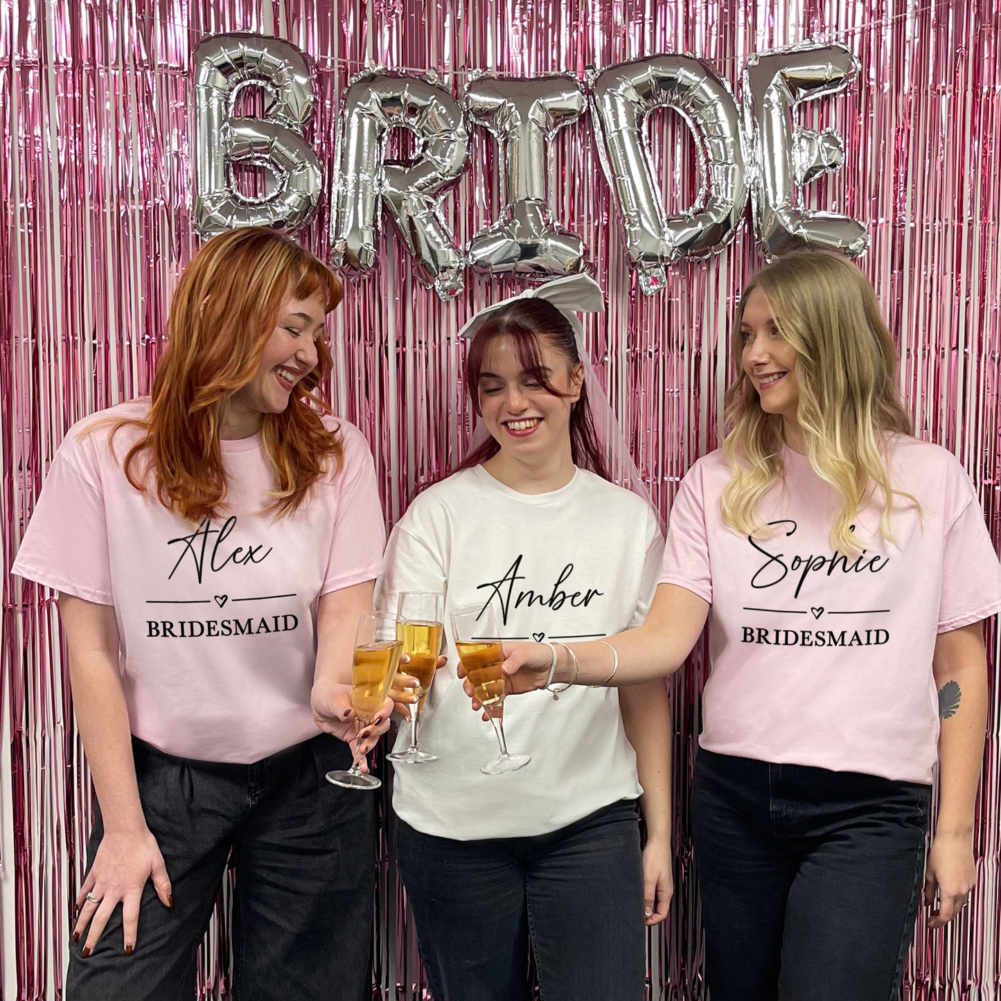 Three women in matching 'Bridesmaid' shirts with personalized names, holding glasses against a pink 'BRIDE' balloon backdrop.