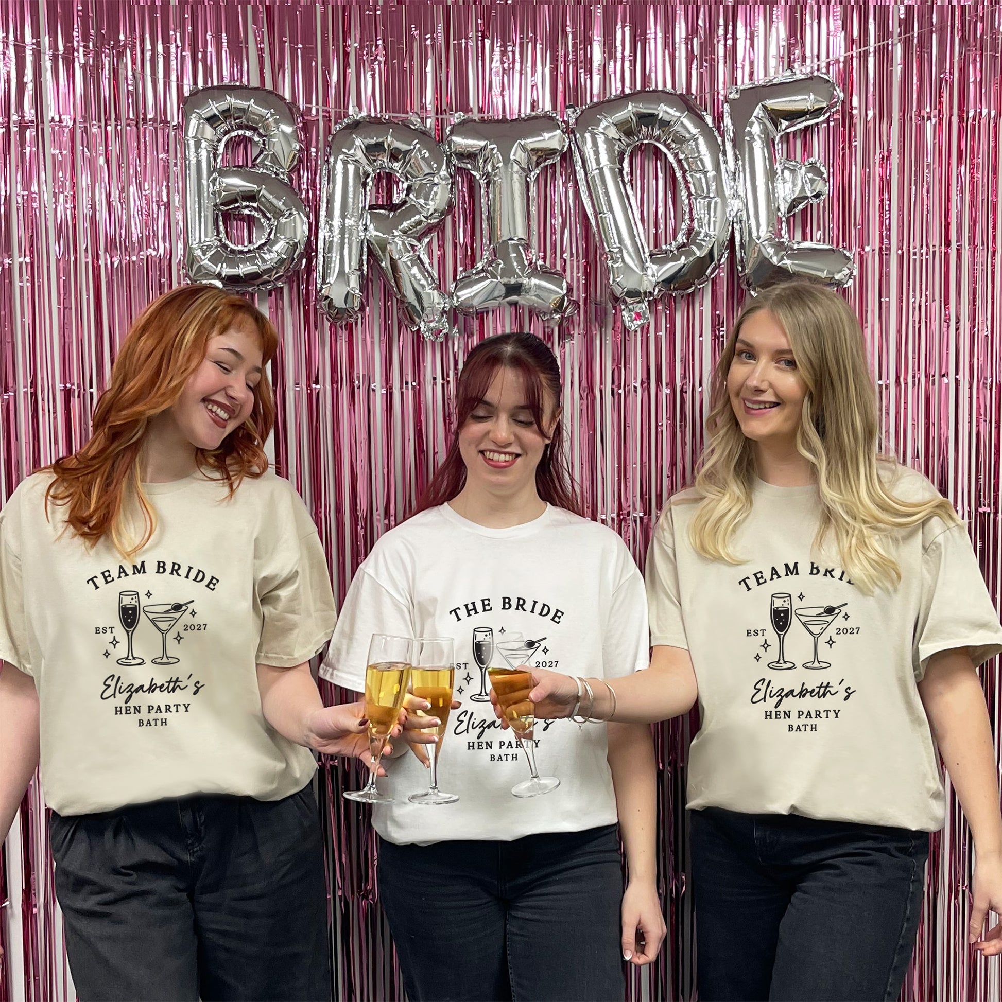 Three women standing together in front of a pink tinsel backdrop with silver foil balloons spelling BRIDE. They are wearing custom printed t-shirts that read 'TEAM BRIDE' and 'Elizabeth's Hen Party 2027' with cocktail graphics.
