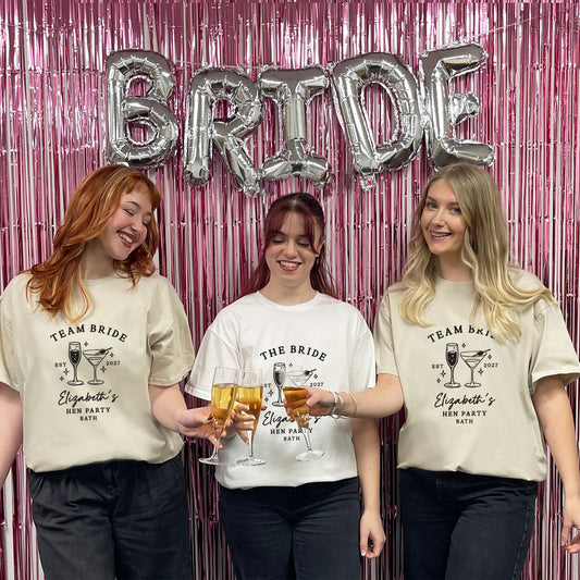 Three women standing together in front of a pink tinsel backdrop with silver foil balloons spelling BRIDE. They are wearing custom printed t-shirts that read 'TEAM BRIDE' and 'Elizabeth's Hen Party 2027' with cocktail graphics.