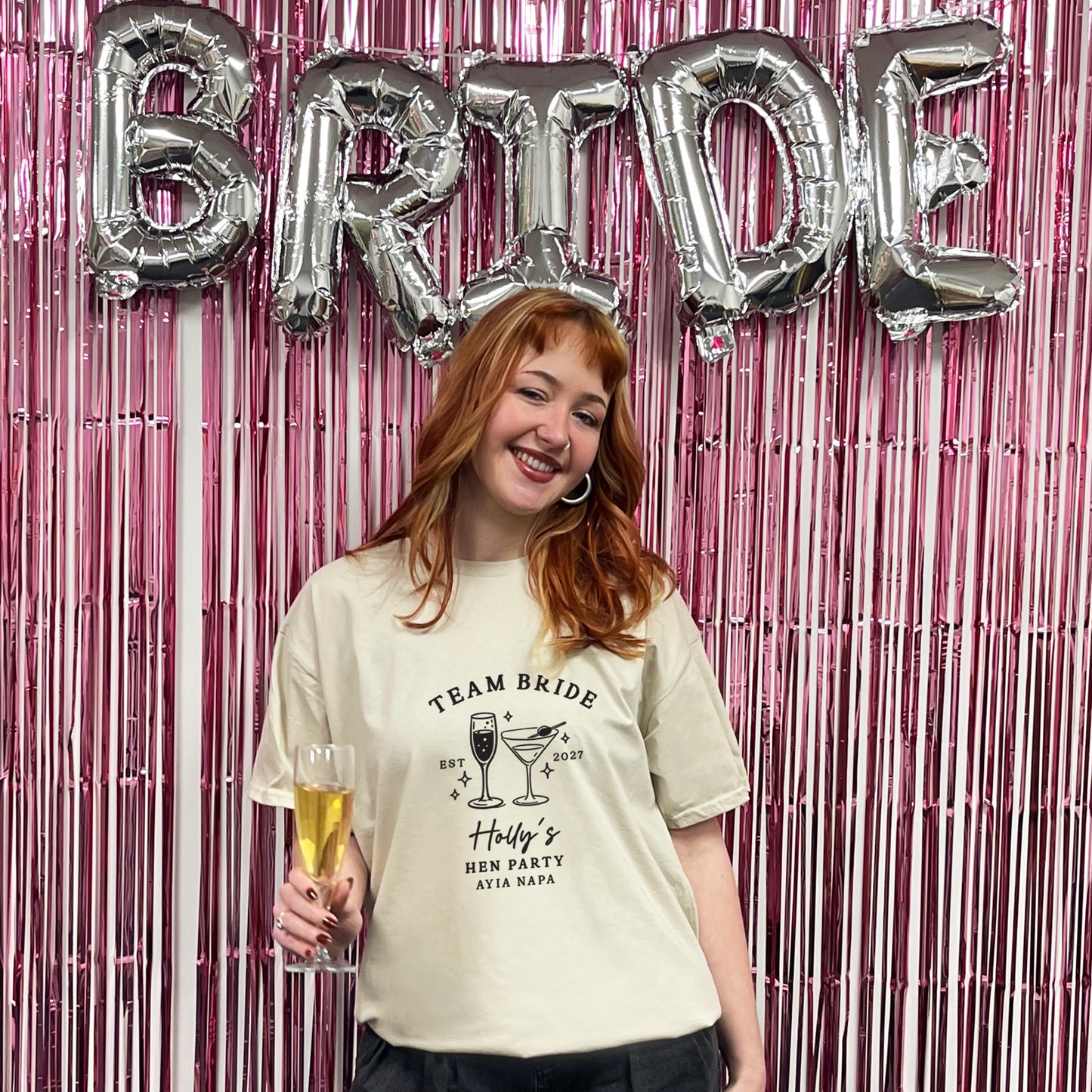A woman wearing a cream-colored graphic t-shirt that reads 'TEAM BRIDE' with cocktail illustrations and 'Holly's Brides Hen Party Atlanta GA' stands in front of a pink metallic fringe backdrop with silver foil 'BRIDE' balloons.