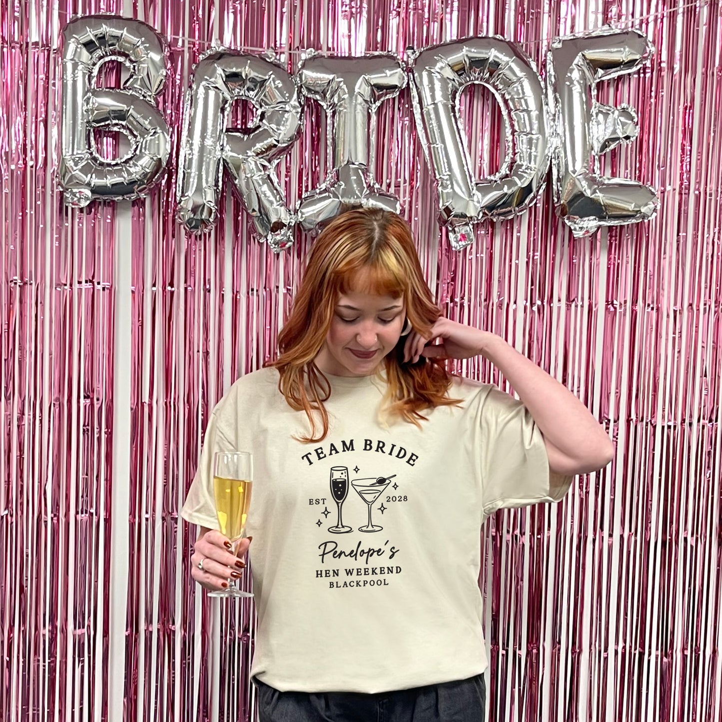 A woman stands in front of a pink metallic tinsel backdrop with large silver foil balloons spelling BRIDE. She is wearing a cream graphic T-shirt that reads 'TEAM BRIDE' and 'Pinaud's Hen Weekend Blackpool,' while holding a glass of champagne.