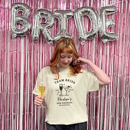 A woman stands in front of a pink metallic tinsel backdrop with large silver foil balloons spelling BRIDE. She is wearing a cream graphic T-shirt that reads 'TEAM BRIDE' and 'Pinaud's Hen Weekend Blackpool,' while holding a glass of champagne.