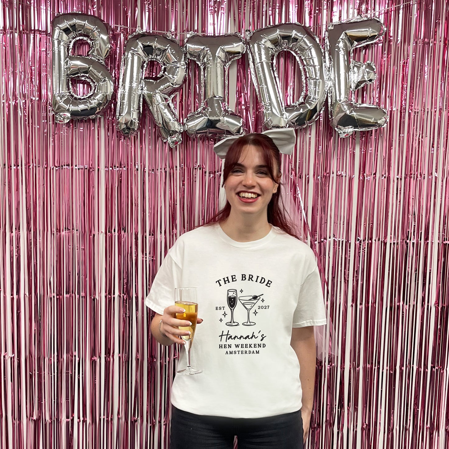 A woman stands in front of a pink metallic fringe backdrop with large silver 'BRIDE' balloons behind her, holding a glass of champagne. She is wearing a white graphic T-shirt that reads 'THE BRIDE' with cocktail illustrations and 'Hannah's BEE WEEKEND AMSTERDAM'.