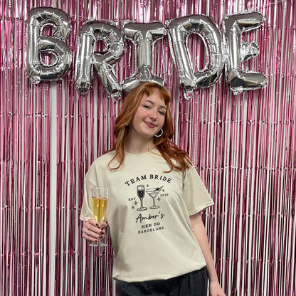 Woman holding a glass of champagne with 'BRIDE' balloons and pink tinsel curtain in the background