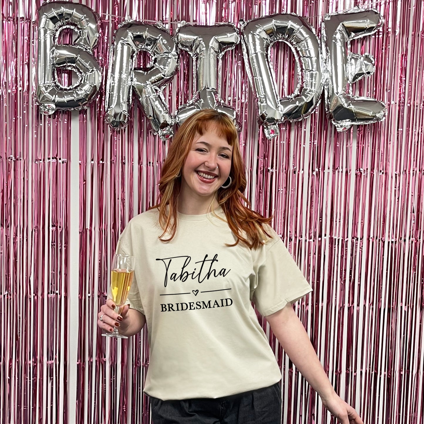 Woman in a 'Bridesmaid' shirt holding a glass of champagne with 'BRIDE' balloons in the background.