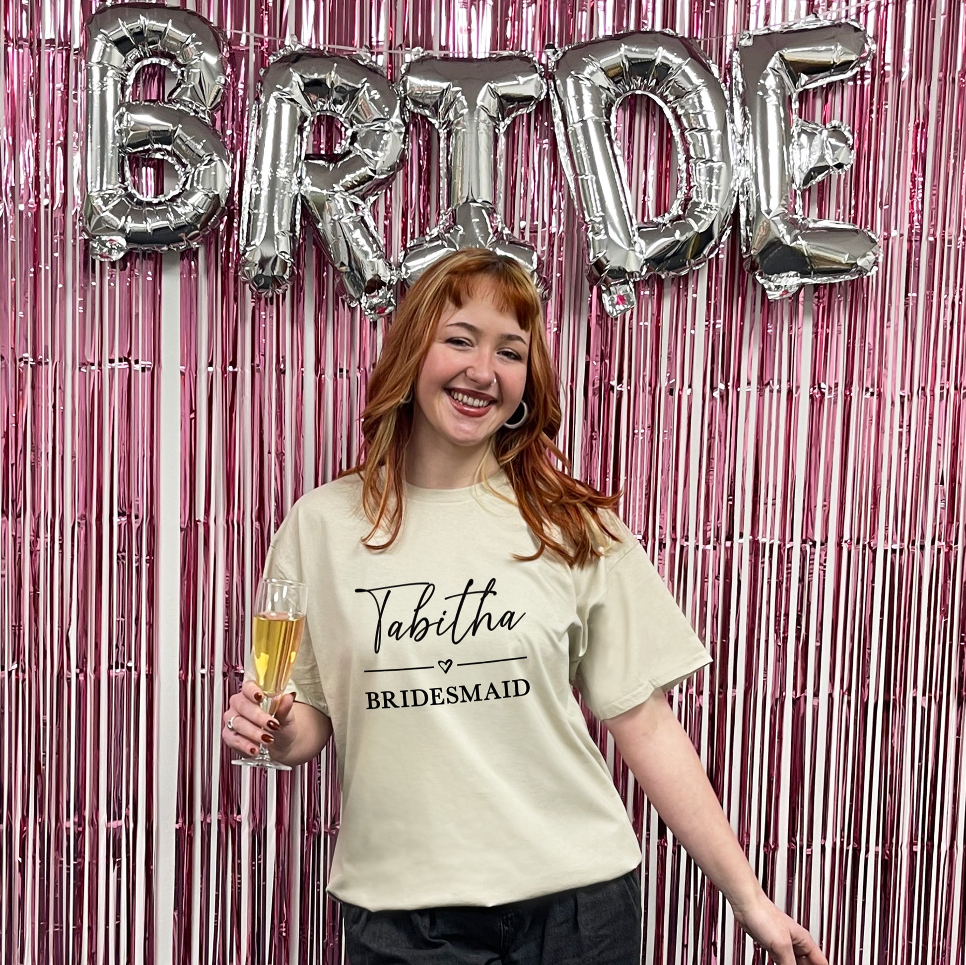 Woman in a 'Bridesmaid' shirt holding a glass of champagne with 'BRIDE' balloons in the background.