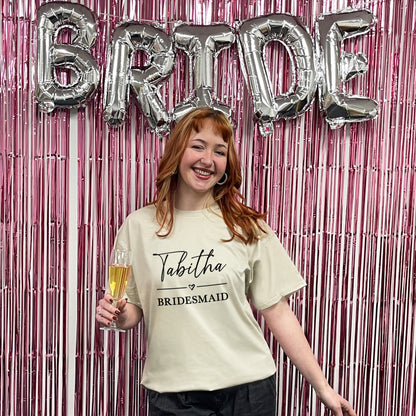 Woman in a 'Bridesmaid' shirt holding a glass of champagne with 'BRIDE' balloons in the background.