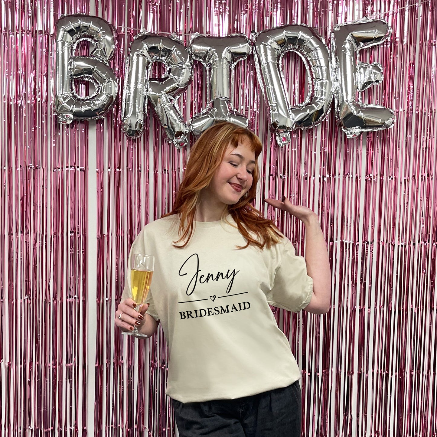 Woman in a 'Jenny Bridesmaid' shirt with 'BRIDE' balloons and a pink fringe curtain.