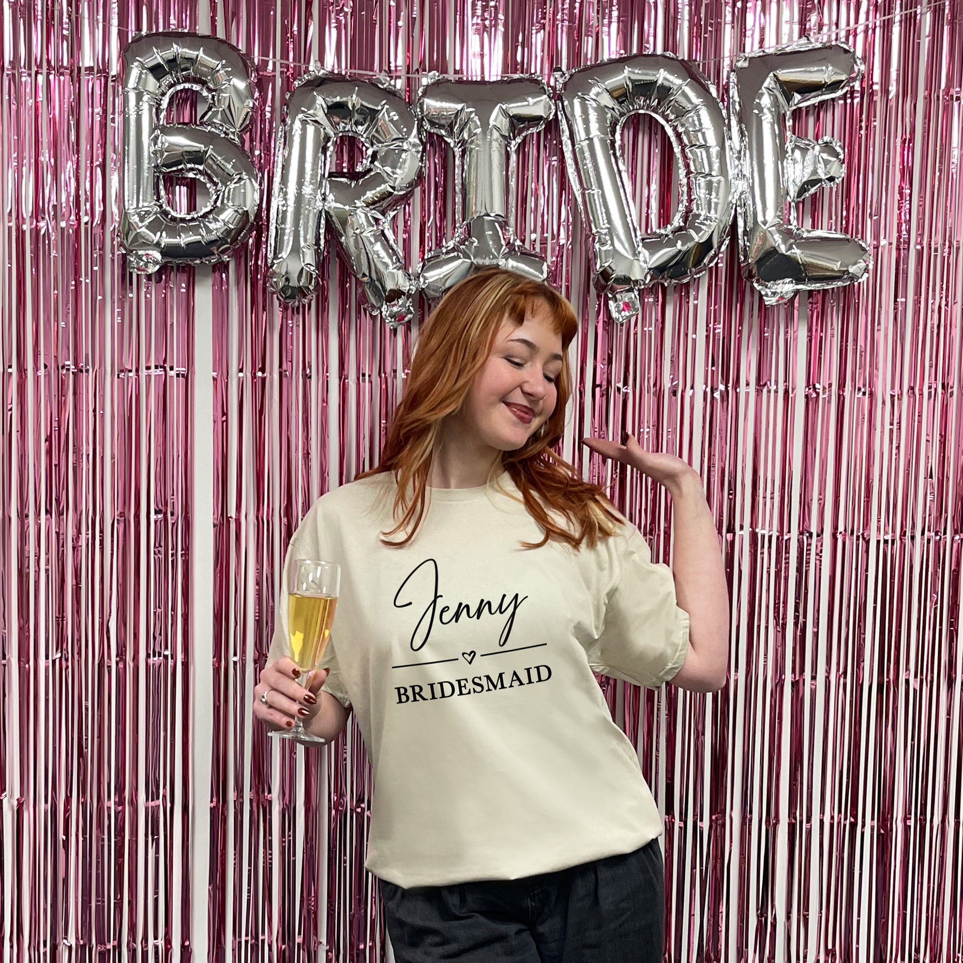 Woman in a 'Jenny Bridesmaid' shirt with 'BRIDE' balloons and a pink fringe curtain.
