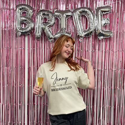 Woman in a 'Jenny Bridesmaid' shirt with 'BRIDE' balloons and a pink fringe curtain.
