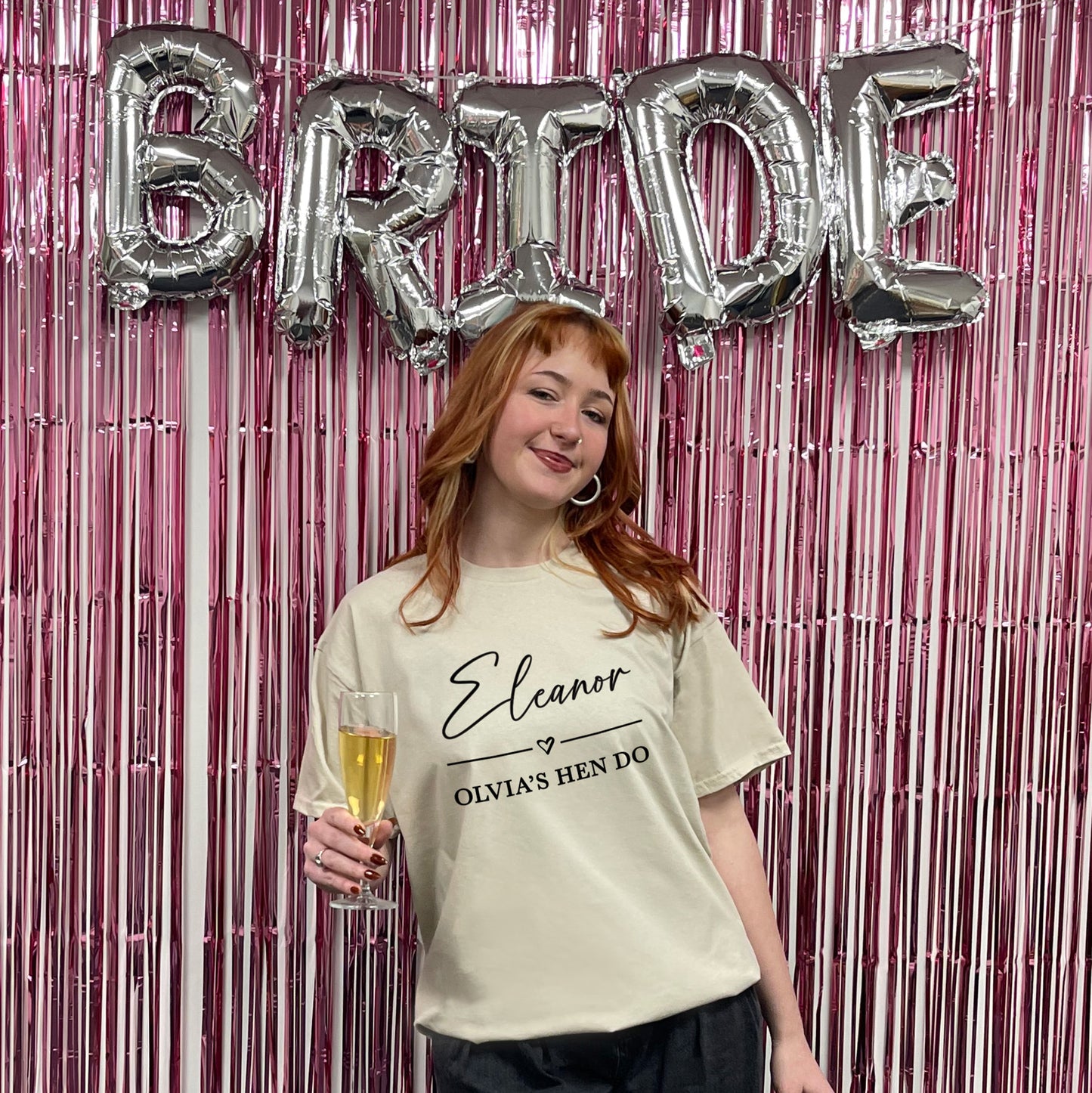 Woman in a 'Joanna Bridesmaid' shirt holding a glass, with 'BRIDE' balloons in the background.