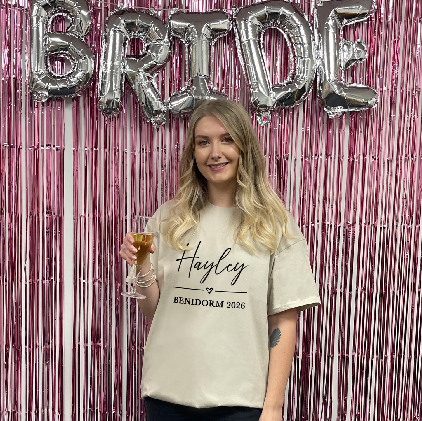 Woman holding a glass of champagne with 'BRIDE' balloons and a pink metallic curtain in the background.