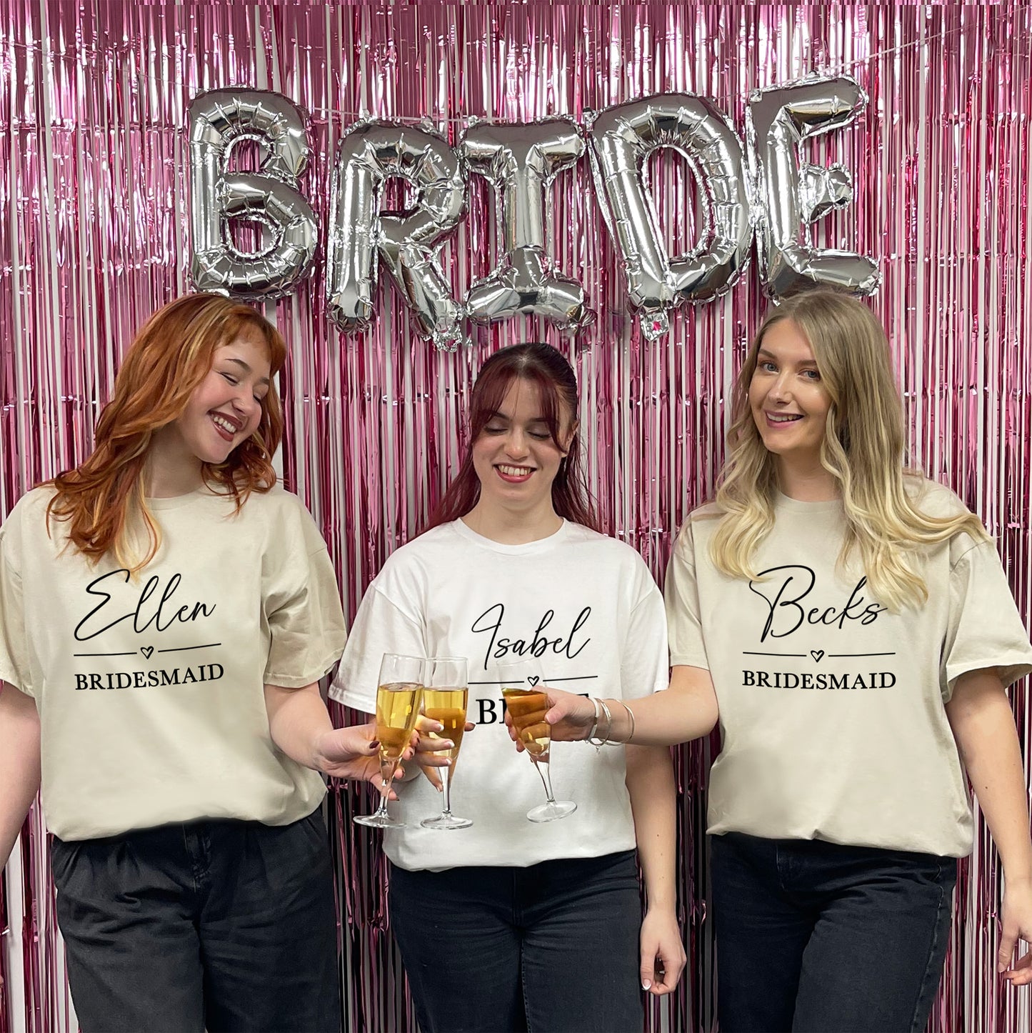 Three women in matching 'Bridesmaid' shirts with personalized names, holding drinks in front of a pink curtain with 'BRIDE' balloons.
