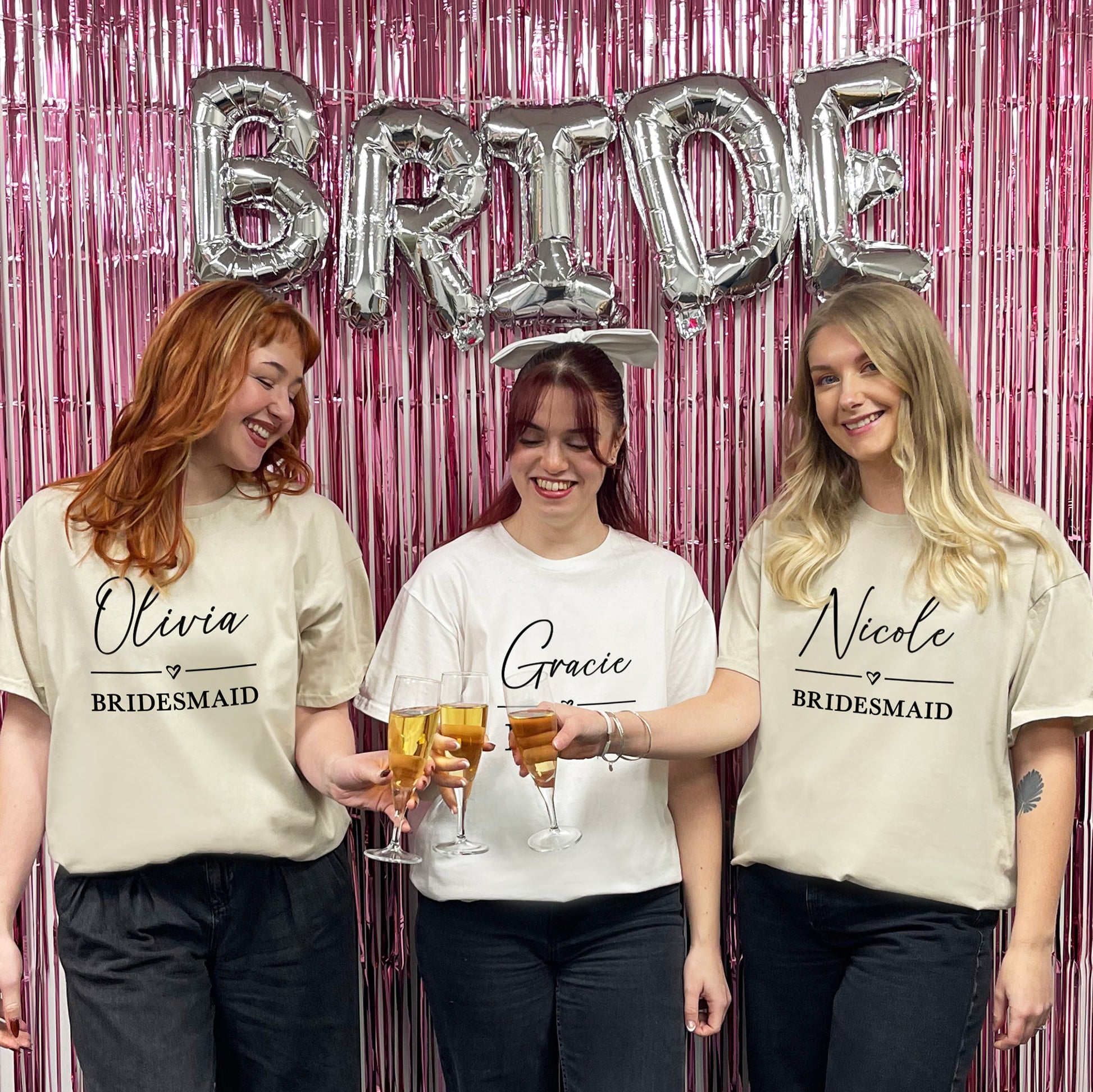 Three women in matching t-shirts with names on, standing in front of a 'BRIDE' balloon and pink fringe curtain.