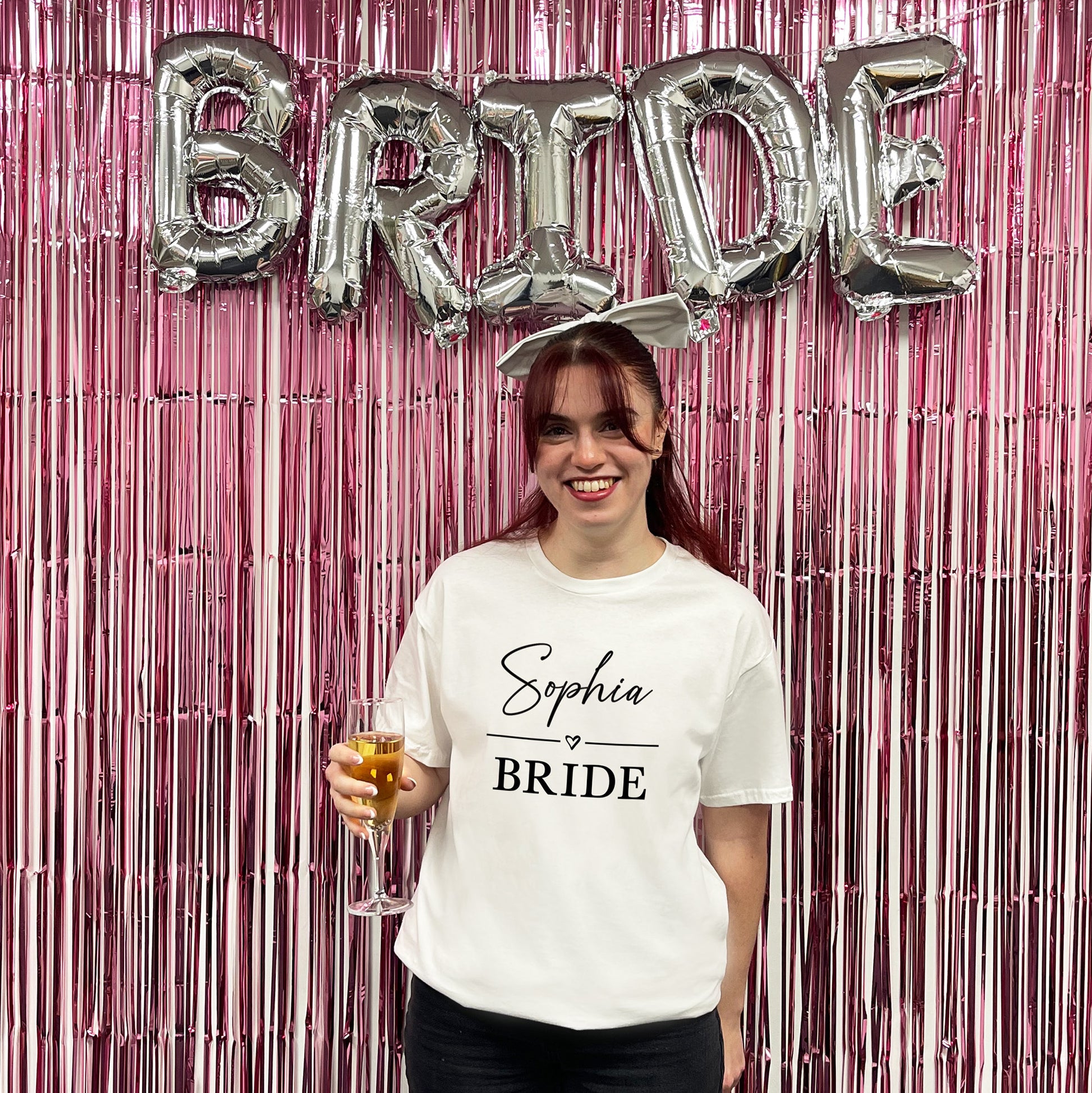 Woman in a 'Sophia Bride' t-shirt with 'BRIDE' balloons and pink streamers in the background.