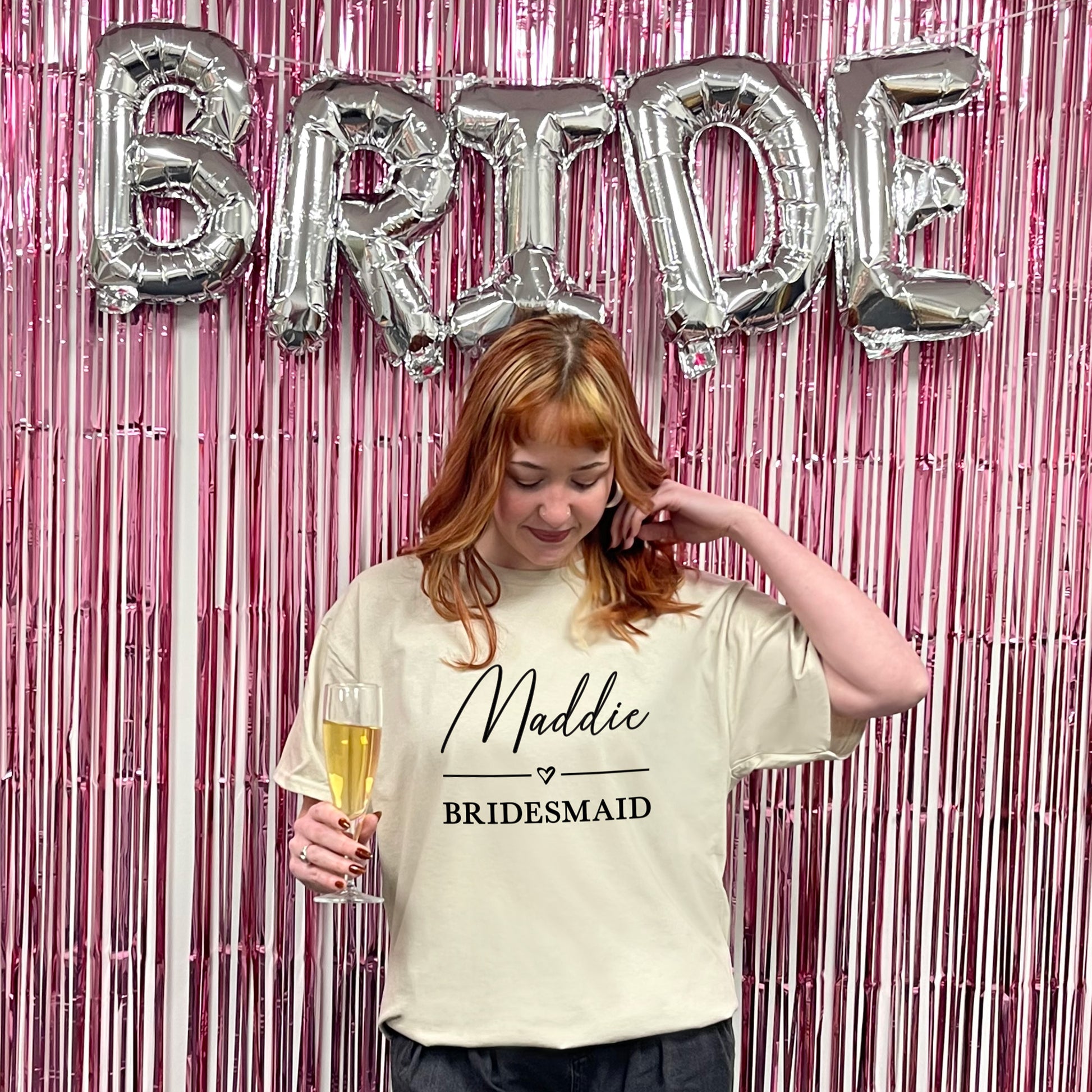 Woman in a 'Maddie Bridesmaid' shirt with 'BRIDE' balloons and pink streamers in the background.