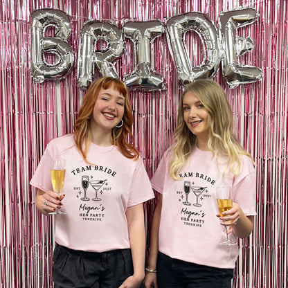 Two women standing in front of a pink tinsel backdrop with silver 'BRIDE' balloons, wearing matching light pink graphic T-shirts that read 'TEAM BRIDE' with cocktail illustrations and text, each holding a champagne flute.