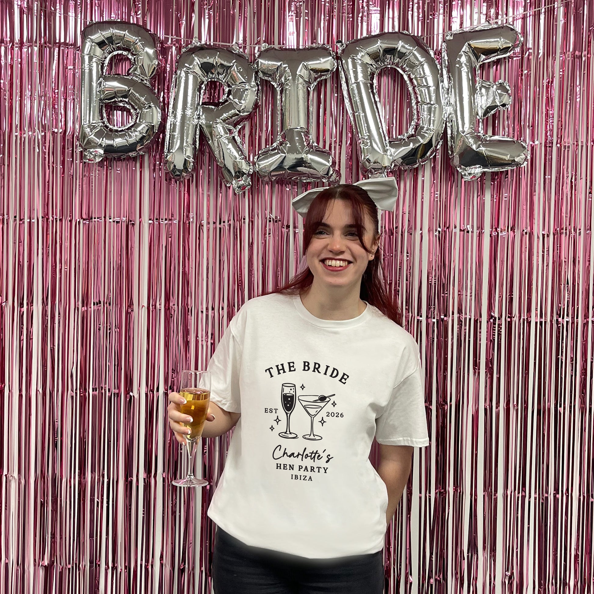 A smiling woman stands in front of a pink metallic fringe backdrop with large silver foil balloons spelling BRIDE. She is wearing a white graphic T-shirt that reads “THE BRIDE” and “Charlotte’s Men Party 1995,” and she holds a drink in a glass.