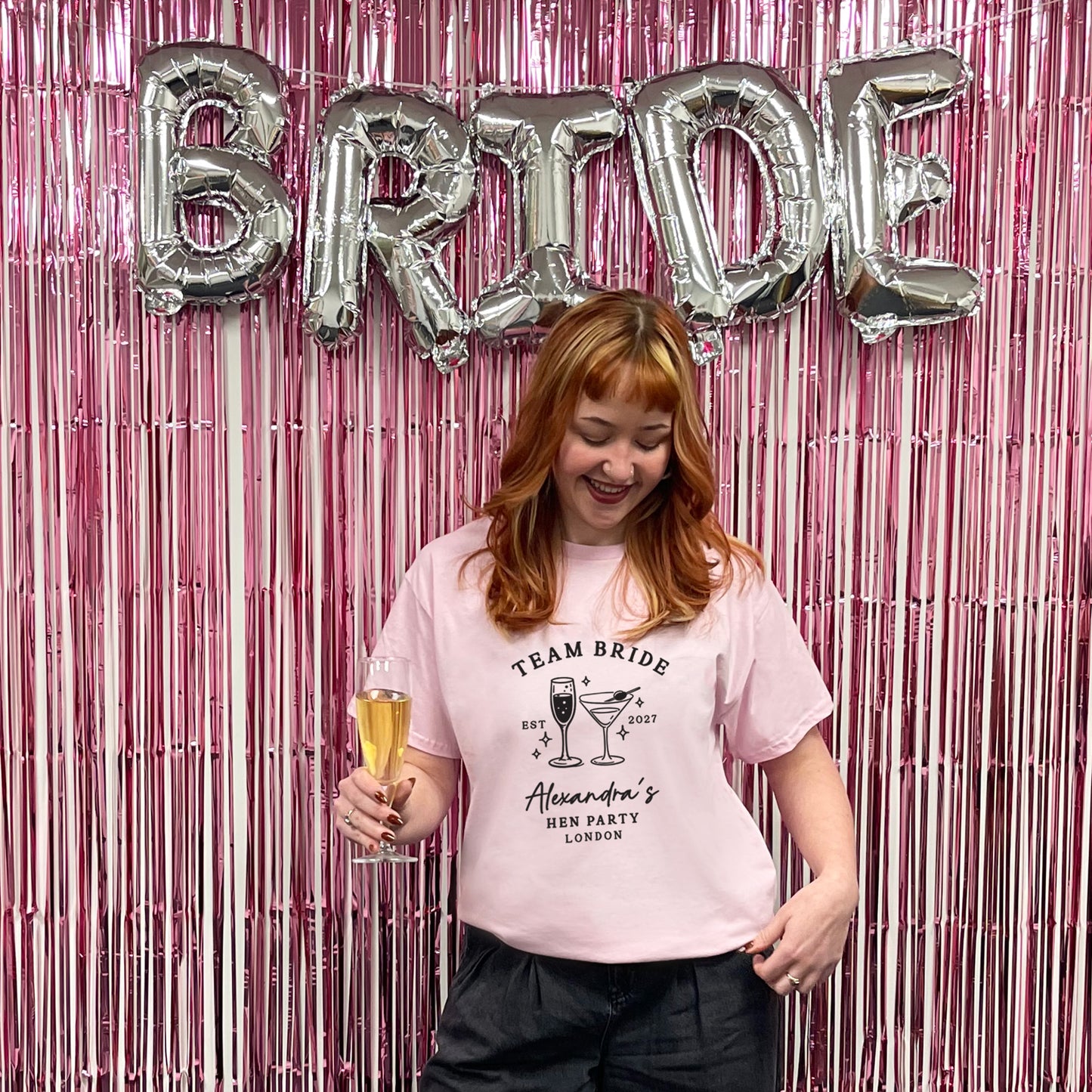 A woman with strawberry-blonde hair wearing a light pink graphic t-shirt that reads 'TEAM BRIDE' and 'Alexander's BEE PARTY LONDON', holding a glass of champagne in front of a pink tinsel backdrop with silver 'BRIDE' balloons.
