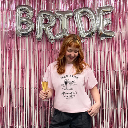 A woman with strawberry-blonde hair wearing a light pink graphic t-shirt that reads 'TEAM BRIDE' and 'Alexander's BEE PARTY LONDON', holding a glass of champagne in front of a pink tinsel backdrop with silver 'BRIDE' balloons.