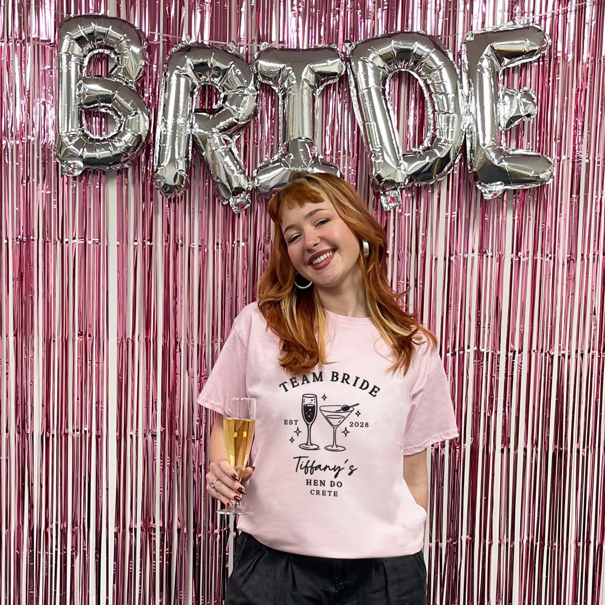 A woman with red hair stands in front of a pink metallic fringe backdrop with large silver 'BRIDE' balloons, wearing a light pink graphic T-shirt that reads 'TEAM BRIDE Tiffany's MEN DO CRISTE' with cocktail illustrations.