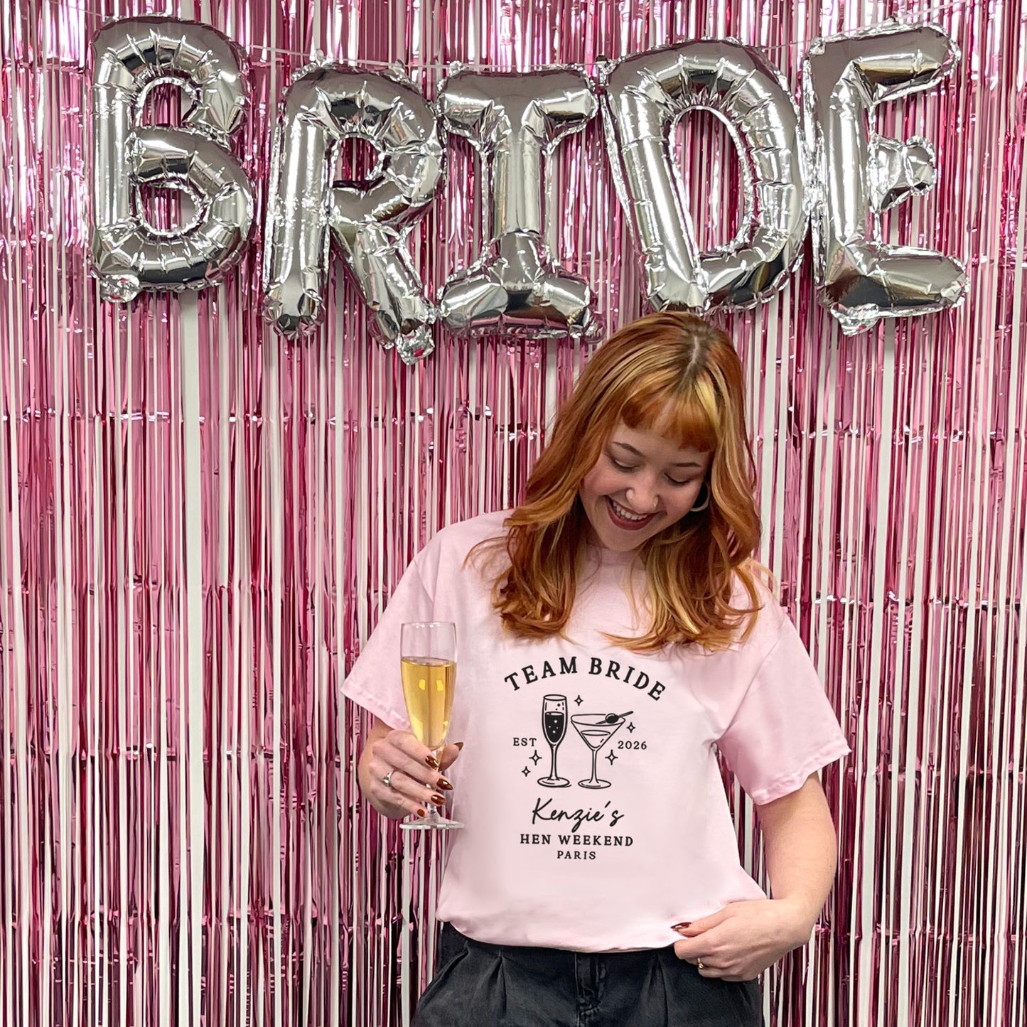 A woman with red hair stands in front of a pink metallic fringe backdrop with large silver foil balloons spelling BRIDE. She is wearing a light pink graphic T-shirt that reads 'TEAM BRIDE' with cocktail illustrations and 'King's Hen Weekend Paris,' while holding a champagne flute.