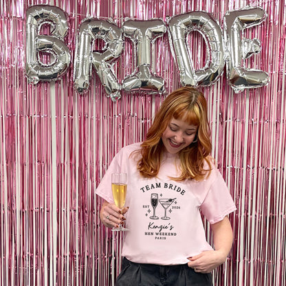 A woman with red hair stands in front of a pink metallic fringe backdrop with large silver foil balloons spelling BRIDE. She is wearing a light pink graphic T-shirt that reads 'TEAM BRIDE' with cocktail illustrations and 'King's Hen Weekend Paris,' while holding a champagne flute.