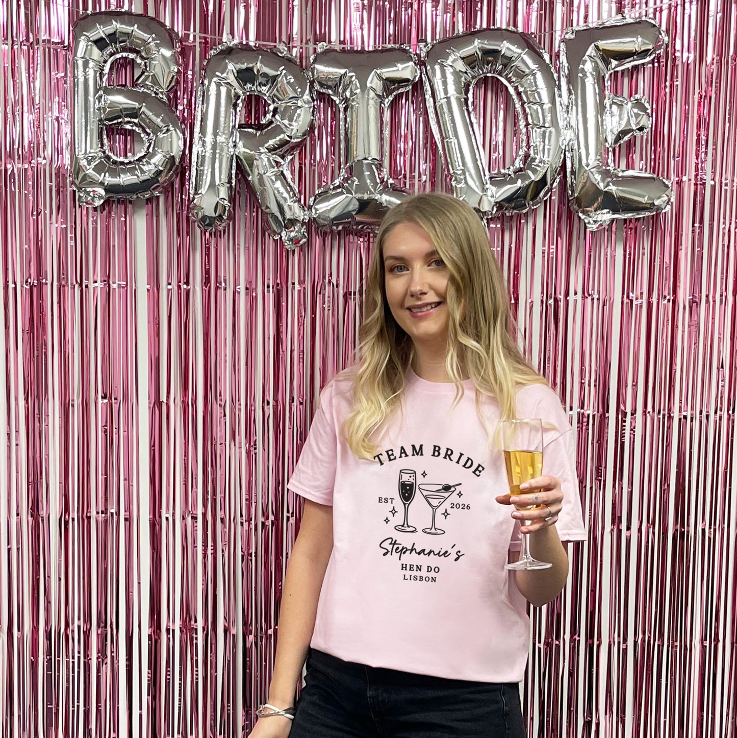 A woman stands in front of a pink metallic fringe backdrop with large silver foil letters spelling BRIDE. She is wearing a light pink graphic T-shirt that reads 'TEAM BRIDE' with cocktail illustrations and the name 'Stephanie's MEN DO LONDON,' and she holds a champagne flute.