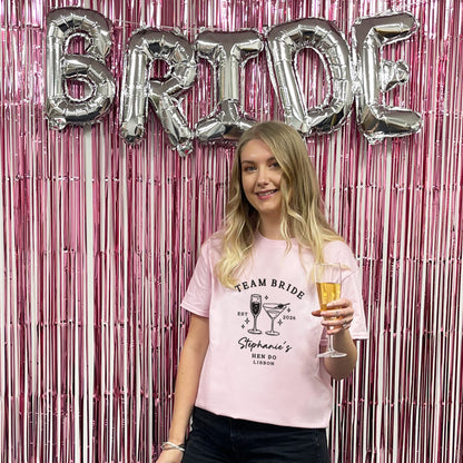 A woman stands in front of a pink metallic fringe backdrop with large silver foil letters spelling BRIDE. She is wearing a light pink graphic T-shirt that reads 'TEAM BRIDE' with cocktail illustrations and the name 'Stephanie's MEN DO LONDON,' and she holds a champagne flute.