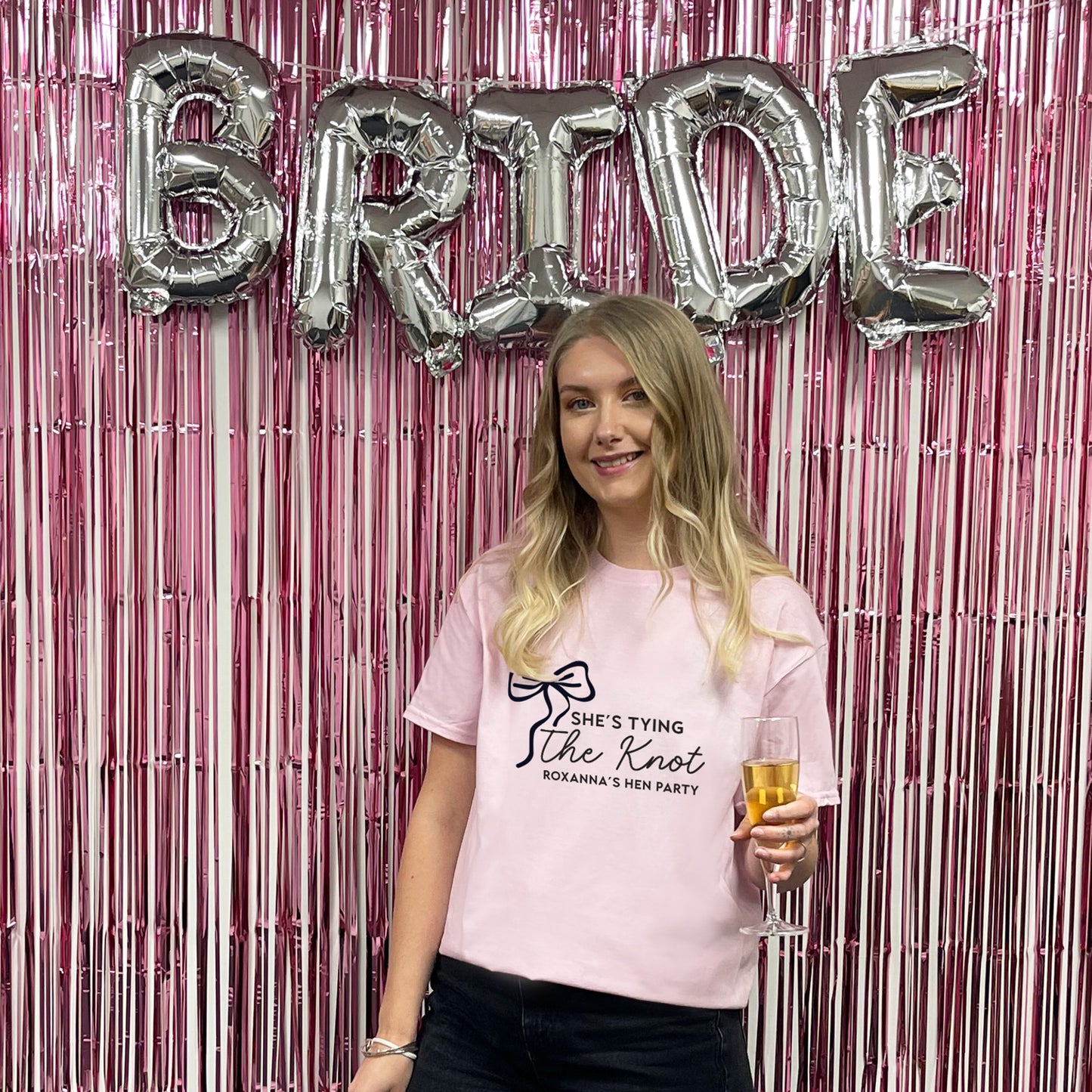 Woman holding a glass of champagne in front of 'BRIDE' balloons and pink tinsel curtain.